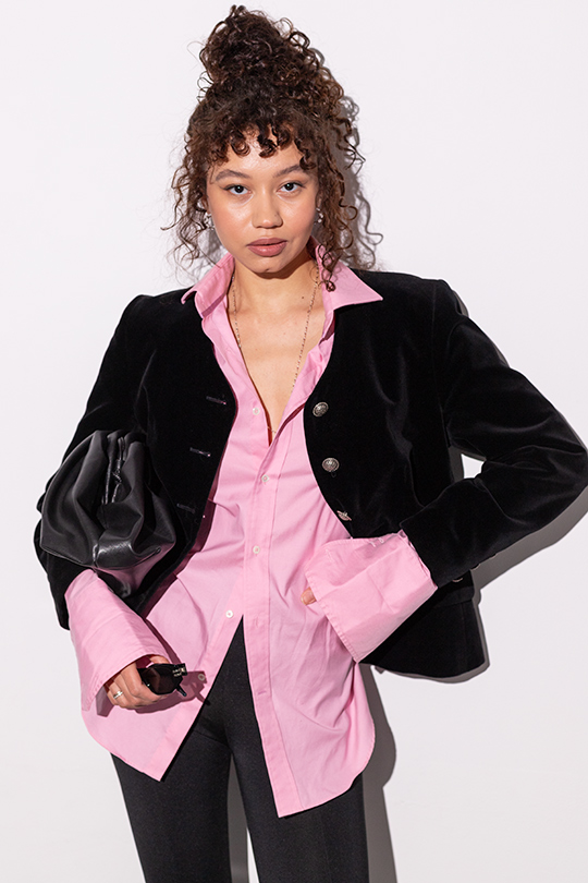 Девушка в розовой рубашке и черном бархатном пиджаке держит под мышкой сумку-баул.